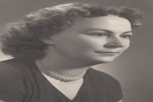 Ein Schwarz-Weiß-Foto einer Frau mit ernstem Gesichtsausdruck, die eine Kette und ein einfaches Kleid mit hohem Kragen und langen Ärmeln trägt, ihre Haare zurückgebunden und ihre Augen konzentriert.