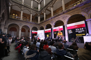Eine Gruppe von Menschen, die auf einer Reihe von Stühlen vor einem Publikum sitzen, wobei einige Kameras halten, während einer Pressekonferenz für das CDMX 2017-Event, mit einem Podium mit einem Mikrofon in der Mitte, Bannern mit Text und Bildern, Säulen, Geländern, Fenstern und einer Wand im Hintergrund.