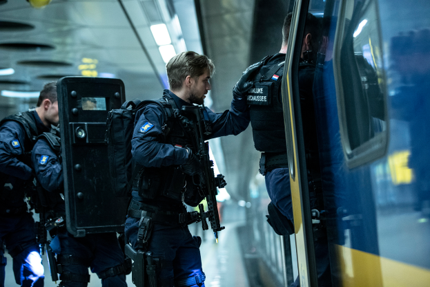 Eine Gruppe von Polizisten steht neben einem Zug auf der rechten Seite des Bildes, wobei einer von ihnen eine Waffe hält und an der Decke befestigte Lampen zu sehen sind.