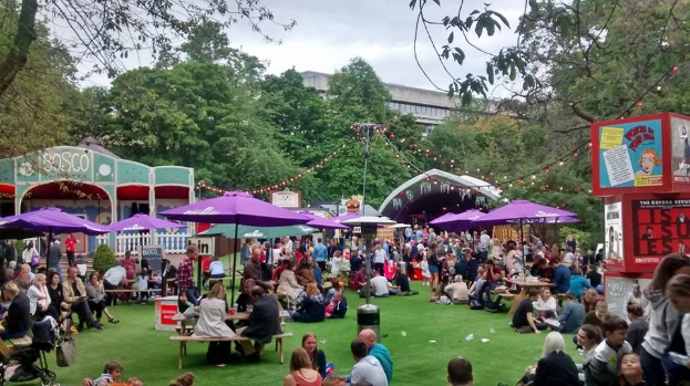 Große Gruppe von Menschen auf einer grünen Wiese mit Schirmen, Tischen und einer Tafel, die ein Top-Biergarten in London anzeigt, umgeben von Bäumen und Gebäuden.