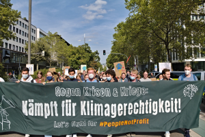 Eine Gruppe maskierter Personen mit einem Banner "Lass uns für die Menschen kämpfen, nicht für den Profit" vor einer belebten Straßenszene mit Fahrzeugen, Bäumen, Gebäuden, Laternen und Verkehrszeichen unter einem bewölkten Himmel.