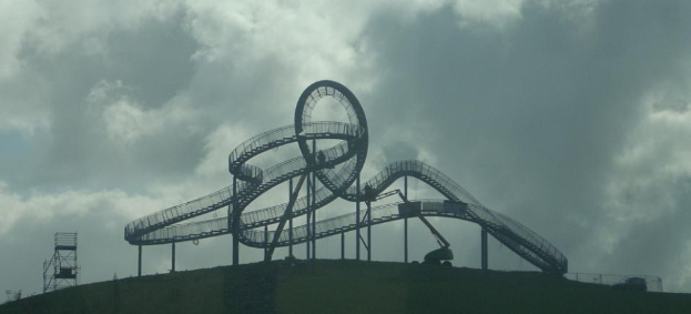 Eine Achterbahn auf dem Höhepunkt eines Hügels unter einem bewölkten Himmel, umgeben von Stützpfählen und -strukturen.