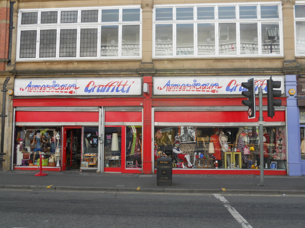 Ein Gebäude mit einem Schild, einem Müllcontainer und einem Pfahl auf einer Straße mit Ampeln, das Puppen und Brillengestelle auf Plakaten zeigt.
