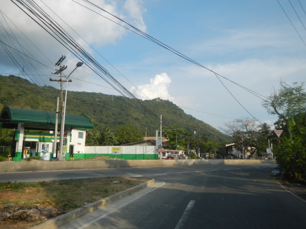 Tankstelle mit elektrischen Masten, Kabeln, Schildern, Bäumen, Gebäuden, Fahrzeugen, Hügeln und bewölktem Himmel im Hintergrund.