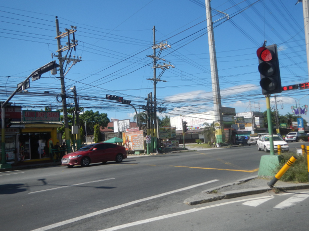 Ein rotes Auto fährt auf einer Straße mit einer Ampel, Strommasten und Kabeln entlang von Gebäuden, Bäumen und einem klaren blauen Himmel im Hintergrund.
