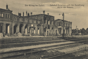Schwarzes und weiße Foto des Bahnhofs Korschen in Ostern, Deutschland, mit Schienensträngen, Gebäuden, Strommasten, Menschen, Bäumen und einem Himmel im Hintergrund.