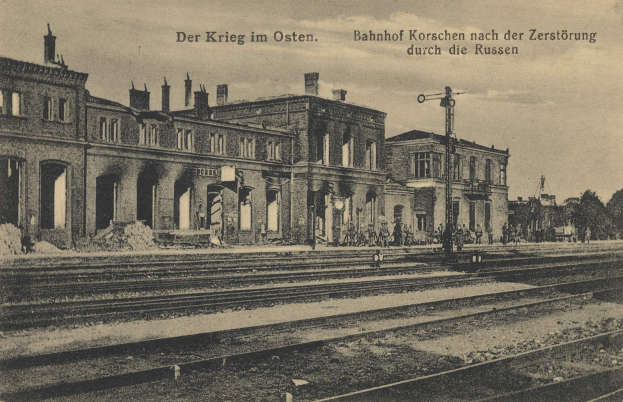 Schwarzes und weiße Foto des Bahnhofs Korschen in Ostern, Deutschland, mit Schienensträngen, Gebäuden, Strommasten, Menschen, Bäumen und einem Himmel im Hintergrund.