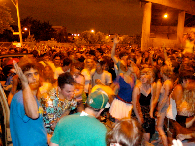 Leute tanzen im Vordergrund auf einem Konzert, mit Lichtern, Polen, Bäumen und anderen Gegenständen im Hintergrund.