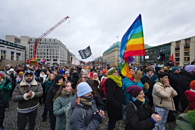 Große Gruppe von Menschen bei einer LGBTQ+-Rechtsdemo in Berlin, mit Fahnen und Plakaten, vor einem Gebäude mit einem Kran und Wolken im Hintergrund.