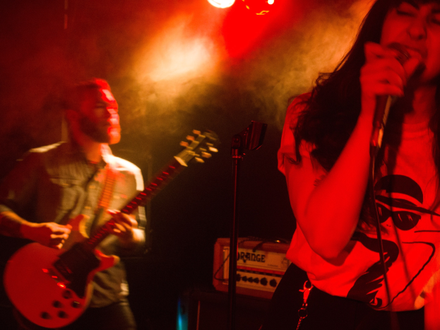 Eine Frau singt in ein Mikrofon auf der Bühne, während ein Mann eine akustische Gitarre im Hintergrund spielt, mit Deckenlampen, die die Szene beleuchten.