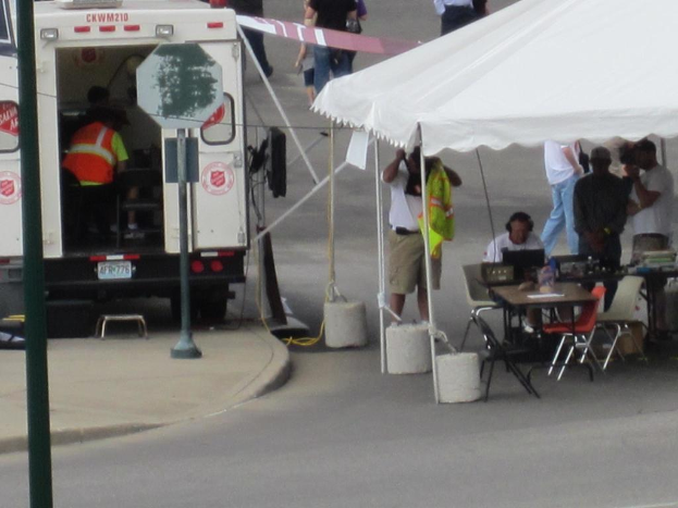 Ein Rettungswagen steht links von einer Stra√üe, w√§hrend auf der rechten Seite ein Zelt eine Gruppe von Menschen sch√ľtzt, mit einem Tisch und einem Stuhl in der N√§he.