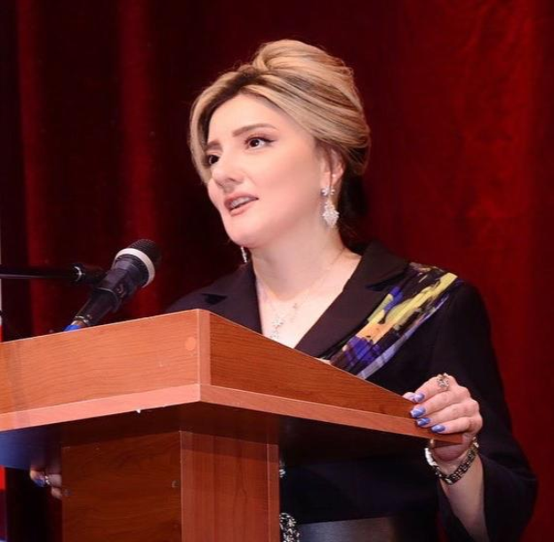 Eine Frau steht an einem Podium mit einem Mikrofon und spricht vor einem roten Vorhang, wahrscheinlich über die Rechte der Frauen.