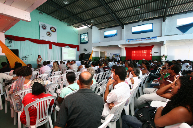 Eine Gruppe von Menschen, die auf Stühlen sitzen und einer Bühne mit einem Polizisten, der in ein Mikrofon spricht, zugewandt sind, wobei auf der rechten Seite der Bildes eine Tür zu sehen ist.