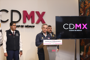 Drei Männer stehen hinter einem Pult vor einem Schild, auf dem "CDMX" steht, mit einer Flagge links und einem Fernseher rechts sowie einer Wand mit Text im Hintergrund.