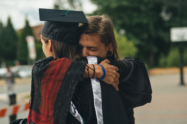 Zwei Personen in Graduationskleidung, die sich gegenseitig umarmen, mit Bäumen im Hintergrund.
