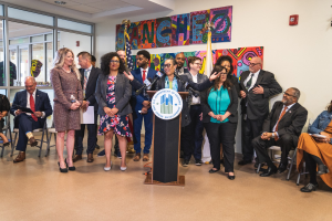 Gruppe von Menschen vor einem Podium mit Mikrofon und einer Tafel mit Text, einige sitzen auf Stühlen und andere stehen, mit Flaggen und einem Feuerlöscher an der Rückwand.