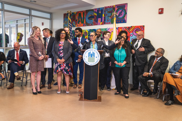 Gruppe von Menschen vor einem Podium mit Mikrofon und einer Tafel mit Text, einige sitzen auf Stühlen und andere stehen, mit Flaggen und einem Feuerlöscher an der Rückwand.