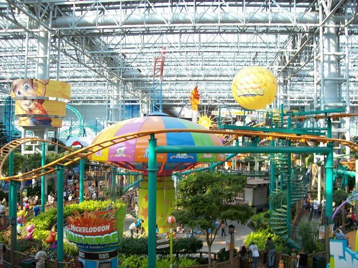 Großes Indoor-Wasserpark mit Achterbahn umgeben von Menschen, Bäumen, Pflanzen und Leuchten an einer farbenfrohen Decke.