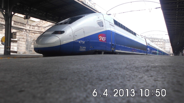 Ein Zug ist in der Mitte des Bildes zu sehen, mit einem Bahnsteig und Text unten, Gebäuden und einem Dach links und Gebäuden, Dach, Kabeln und Himmel oben.