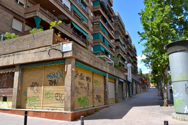 Eine Szene mit Gebäuden, die Fenster, grüne Kleidungsstücke, Geschäfte, Schilder und umgebende Bäume und Pflanzen zeigen.