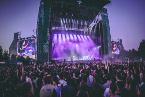 Eine große Menschenmenge auf einem Musikfestival, mit einer Bühne in der Mitte, auf der Musiker Instrumente spielen, beleuchtet von Lichtern und umgeben von Bildschirmen, mit einem Gebäude, Bäumen und einem klaren blauen Himmel im Hintergrund.