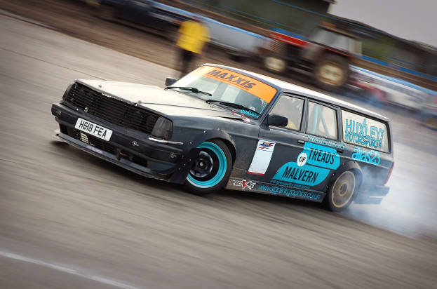 Ein Auto fährt eine Rennstrecke entlang und produziert Rauch, umgeben von anderen Fahrzeugen und Menschen.