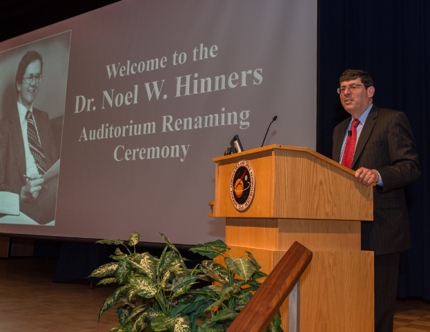Ein Mann in Blazer, Hose und Krawatte steht hinter einem Podium mit einem Mikrofon, rechts von einer Pflanze und einem Bildschirm mit einem Mann und Text.