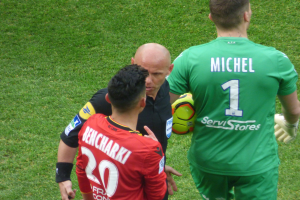 Schiedsrichter spricht mit zwei Fußballspielern auf einem saftig grünen Feld, ein Spieler hält einen Ball.