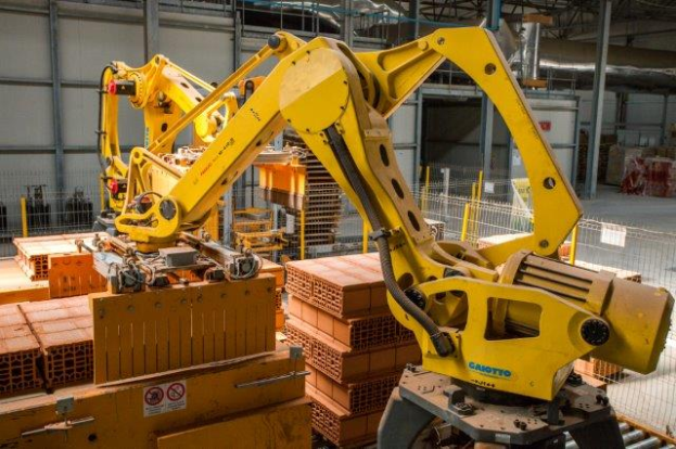 Ein großes Lager mit zahlreichen Kisten, Kränen, einem Zaun, Polen und anderem industriellen Equipment, mit einer Wand im Hintergrund, die auf eine größere industrielle Einrichtung hinweist.