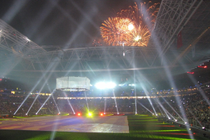 Hell erleuchtetes Olympisches Stadion in London bei Nacht, umgeben von einer großen Menge, mit grünem Gras und Feuerwerk, das den Himmel erhellt.
