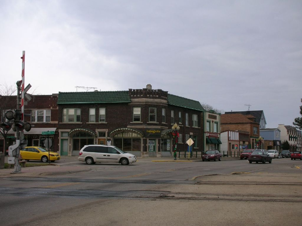 Mehrere Autos fahren auf einer Straße mit Gebäuden mit Fenstern an den Seiten, Ampeln und einem an Pfosten angebrachten Schild sowie einem Baum in der rechten Ecke.