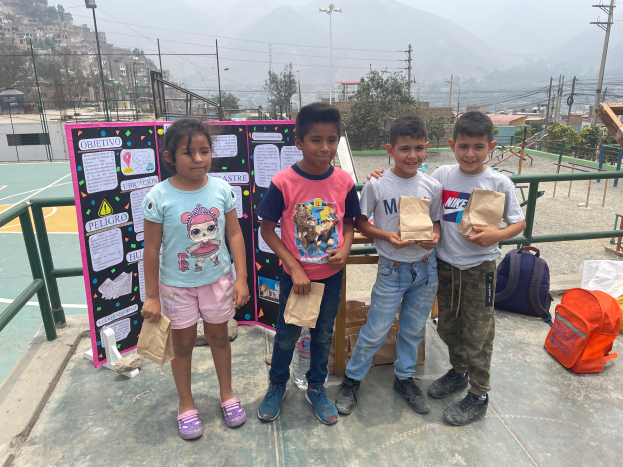 Eine Gruppe von Kindern steht auf einem Basketballfeld, jedes Kind hält eine Papiertüte in der Hand, mit Tüten und Gegenständen rechts auf dem Boden und einer Tafel mit Text links, mit Zaun, Pfosten, Straßenlaternen, Strommasten mit Drähten, Bäumen, Gebäuden, Hügeln und Himmel im Hintergrund.