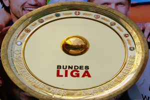 Goldener Schild mit "Bundesliga"-Text, gehalten von einem Mann, mit einem Banner im Hintergrund, das drei Personen zeigt.