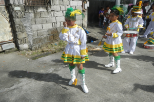 Gruppe junger Mädchen in weißen und gelben Outfits marschiert die Straße entlang und spielt Musikinstrumente, umgeben von einer Wand, einem Tor und Bäumen.