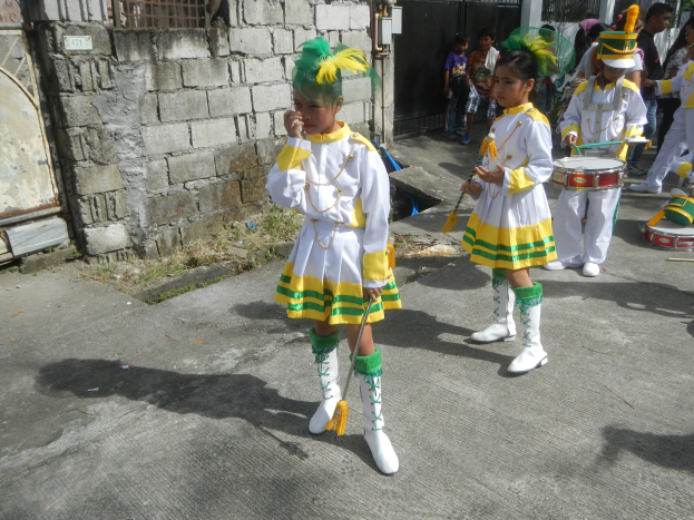 Gruppe junger Mädchen in weißen und gelben Outfits marschiert die Straße entlang und spielt Musikinstrumente, umgeben von einer Wand, einem Tor und Bäumen.