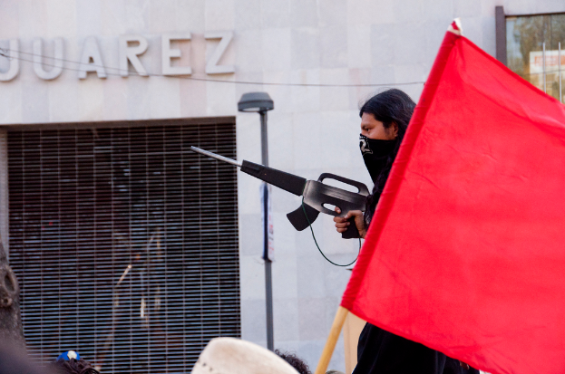 Ein Mann in einem schwarzen Kleid hält eine Waffe, mit einer roten Fahne rechts daneben, eine Straßenlaterne in der Nähe einer Wand und eines Gebäudes im Hintergrund und lesbarer Text an der Wand in der oberen linken Ecke.