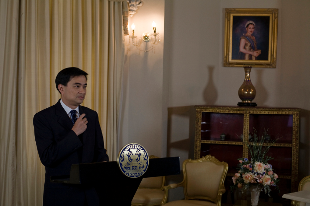 Eine Person steht an einem Rednerpult mit einem Vorhang dahinter, einer Vase und Stühlen rechts und gerahmten Bildern mit daran befestigten Lampen an der Wand.