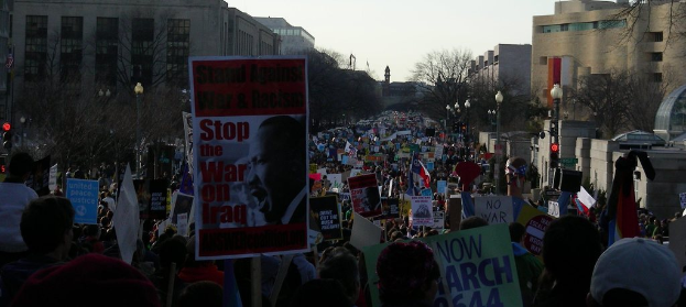 Menschen auf einer Stra√üe protestierend mit Plakaten, mit Geb√§uden, B√§umen, Laternenm√§sten und dem Himmel im Hintergrund.