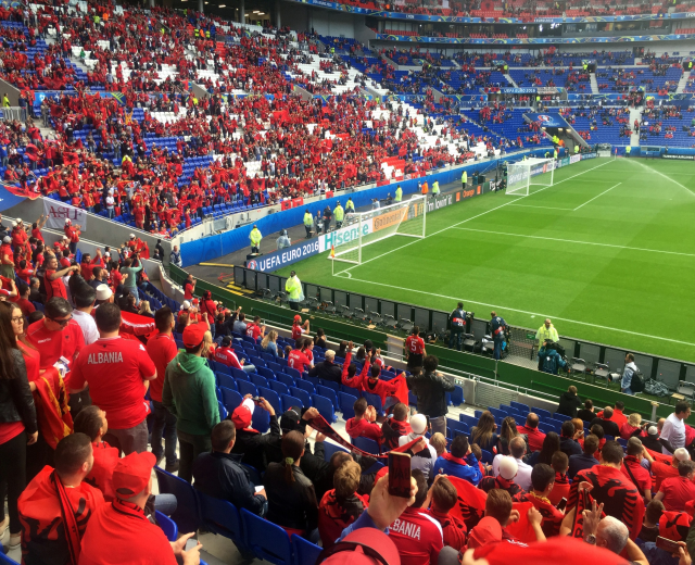 Eine große Menschenmenge in einem Stadion schaut einem Fußballspiel zu, mit Menschen auf Stühlen und Stehplätzen, Transparenten, einem Torpfosten und Deckenleuchten.