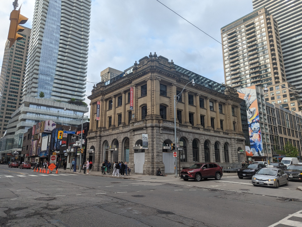 Eine belebte Stadtstraße mit hohen Gebäuden, Fahrzeugen, Fußgängern, Verkehrselementen, Bäumen und der Toronto Public Library als Mittelpunkt unter einem bewölkten Himmel.