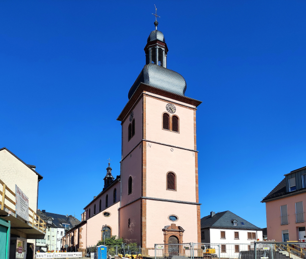 Die Kirche Unserer Lieben Frau der Aufnahme in Baden-Württemberg, Deutschland, ein großes rosafarbenes Gebäude mit einem zentralen Uhrenturm, umgeben von einem Metallzaun, mit benachbarten Gebäuden, Fahrzeugen, Pflanzen, einem Schild, einer Fahne mit Text, Pfählen und einem bewölkten Himmel.