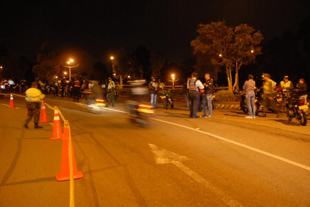 Männer und Fahrräder sind auf einer Straße mit orangefarbenen Fahrbahnbegrenzungen auf der linken Seite, Bäume und Lichter im Hintergrund.
