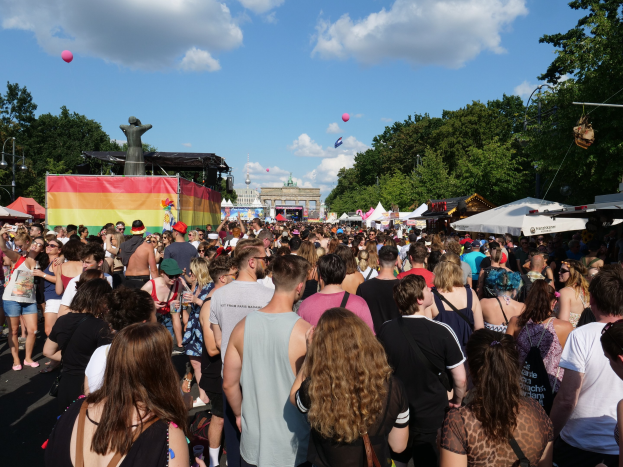 Eine große Menge Menschen geht eine Straße entlang, gesäumt von Zelten, Bäumen, Pfählen, Lichtern und einer Statue bei der Berlin Pride Parade, mit Gebäuden und einem bewölkten Himmel mit Ballons im Hintergrund.