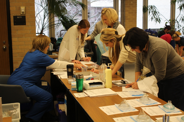 Ein Raum mit Menschen, die in der Nähe eines Schreibtisches mit Papieren, Büchern und Flaschen stehen, eine Frau in einem blauen Kleid in der Ecke sitzt und große Glasfenster mit Pflanzen im Hintergrund