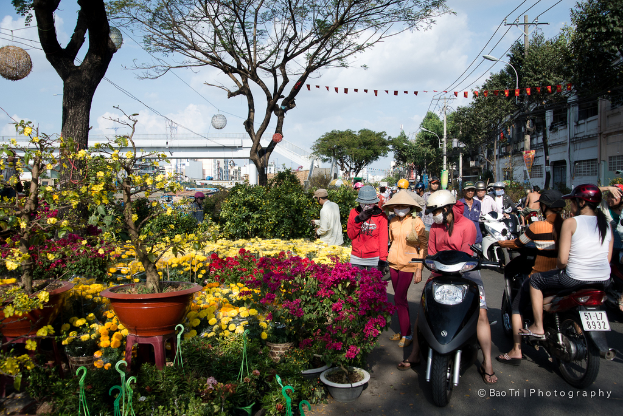 Eine Gruppe von Menschen mit Helmen und M√∂tzen, einige zu Fu√ü und einige mit Fahrr√°dern auf einer Stra√üe, mit Pflanzen, einem Blumentopf auf einem Hocker, B√§umen, einem Pfahl, Dr√°hten, einer Laterne, einer Br√∞cke und Geb√°uden in der N√§he.