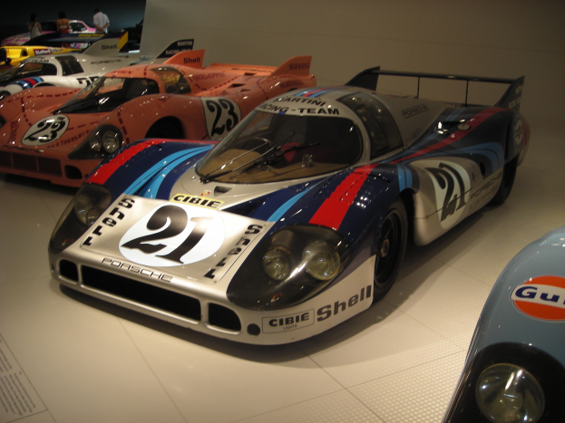 Ein Porsche 917K Le Mans Rennwagen, der in einem Museum ausgestellt ist, umgeben von anderen Sportwagen auf dem Boden. Es gibt Menschen in der Nähe und eine Wand im Hintergrund. Links im Bild gibt es ein Papier mit Text darauf.