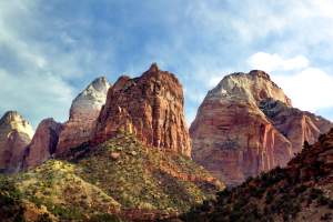 Ein malerischer Blick auf den Zion-Nationalpark in Utah, der majestätische Berge, grüne Bäume, felsiges Gelände und einen Himmel mit weißen, flauschigen Wolken zeigt.