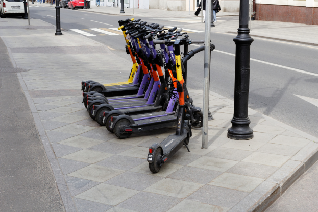 Eine Reihe von Elektro-Scootern, die an einer Straße mit fahrenden Fahrzeugen, Fußgängern auf dem Gehweg und Gebäuden im Hintergrund geparkt sind.