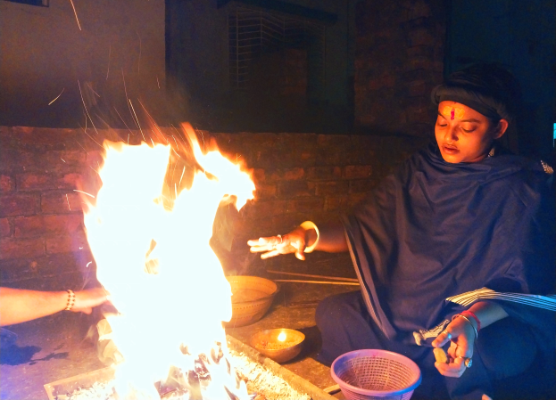 Eine Frau sitzt vor einem Feuer mit ausgestreckten Händen, umgeben von Schüsseln und anderen Gegenständen auf dem Boden, mit einer Wand und einem Fenster im Hintergrund.