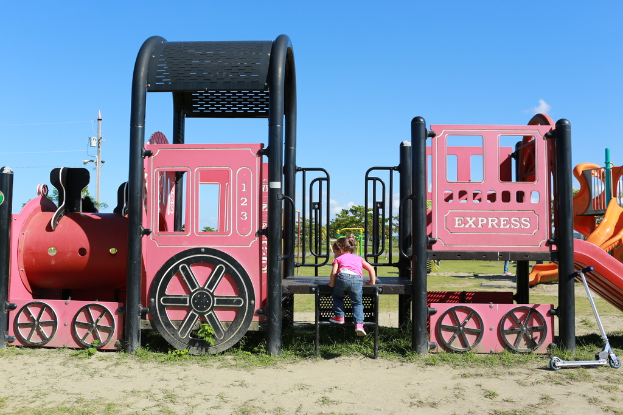 Ein Kind klettert Treppen an Spielplatzgeräten mit Bäumen, Polen, Stromleitungen und Wolken im Hintergrund und einem Roller auf dem Boden rechts.